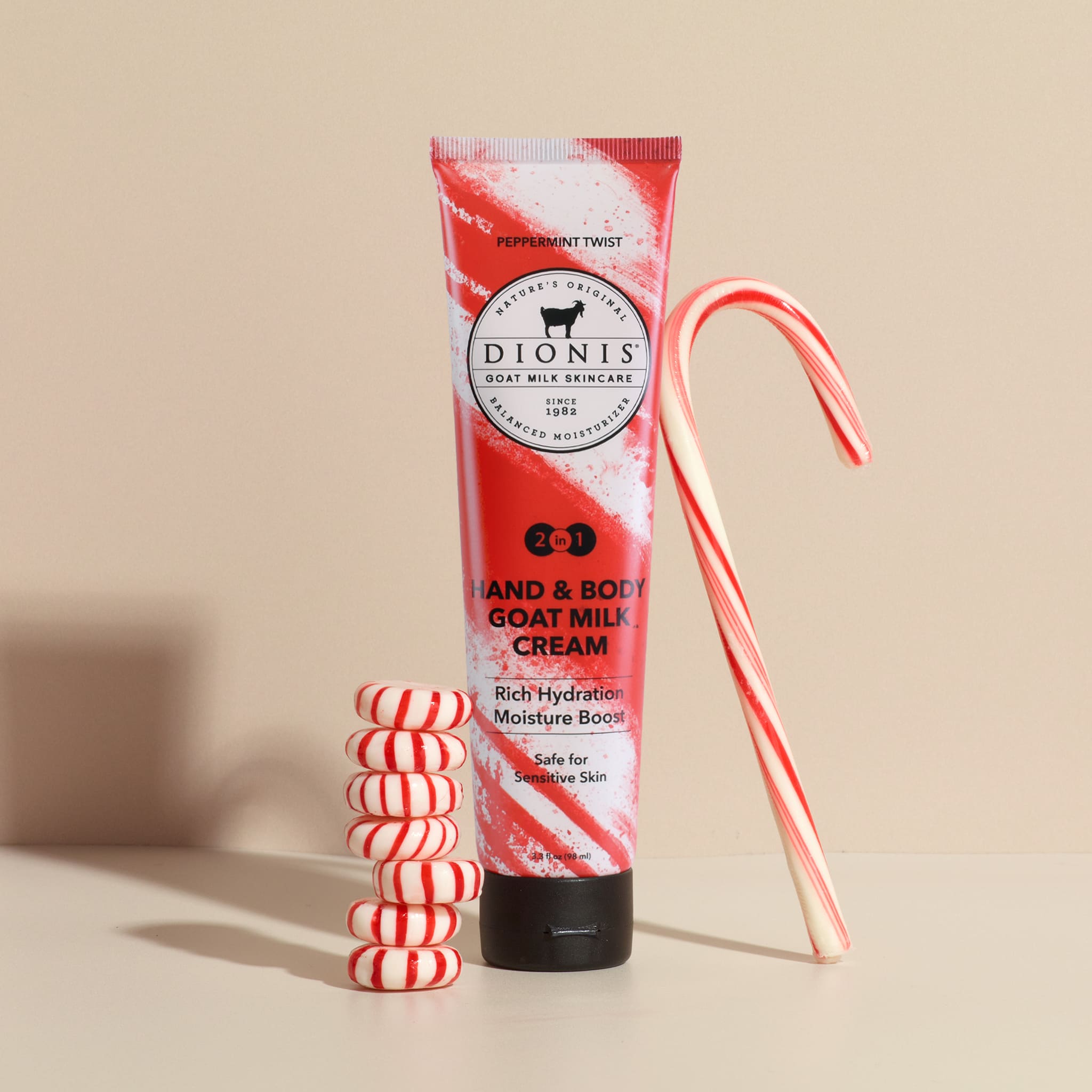A tube of Dionis Hand & Body Goat Milk Cream with a red and white peppermint swirl design, styled with candy canes and stacked peppermint candies on a beige background.