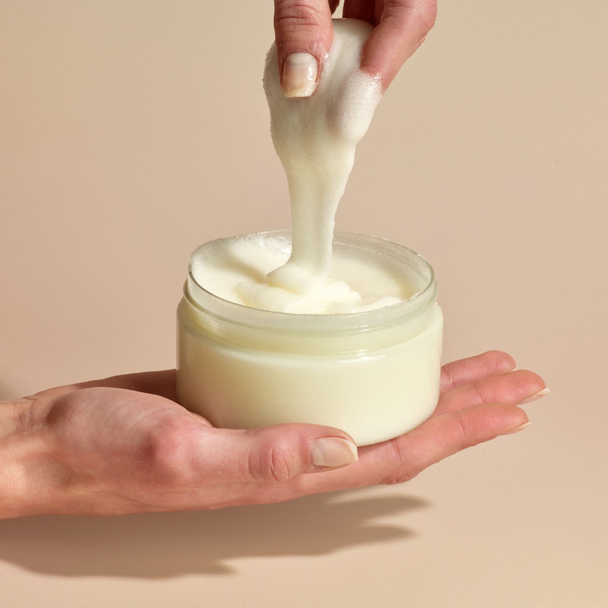 One hand holding Dionis Goat Milk Body Scrub jar while other hand dips in to pull product out and show texture