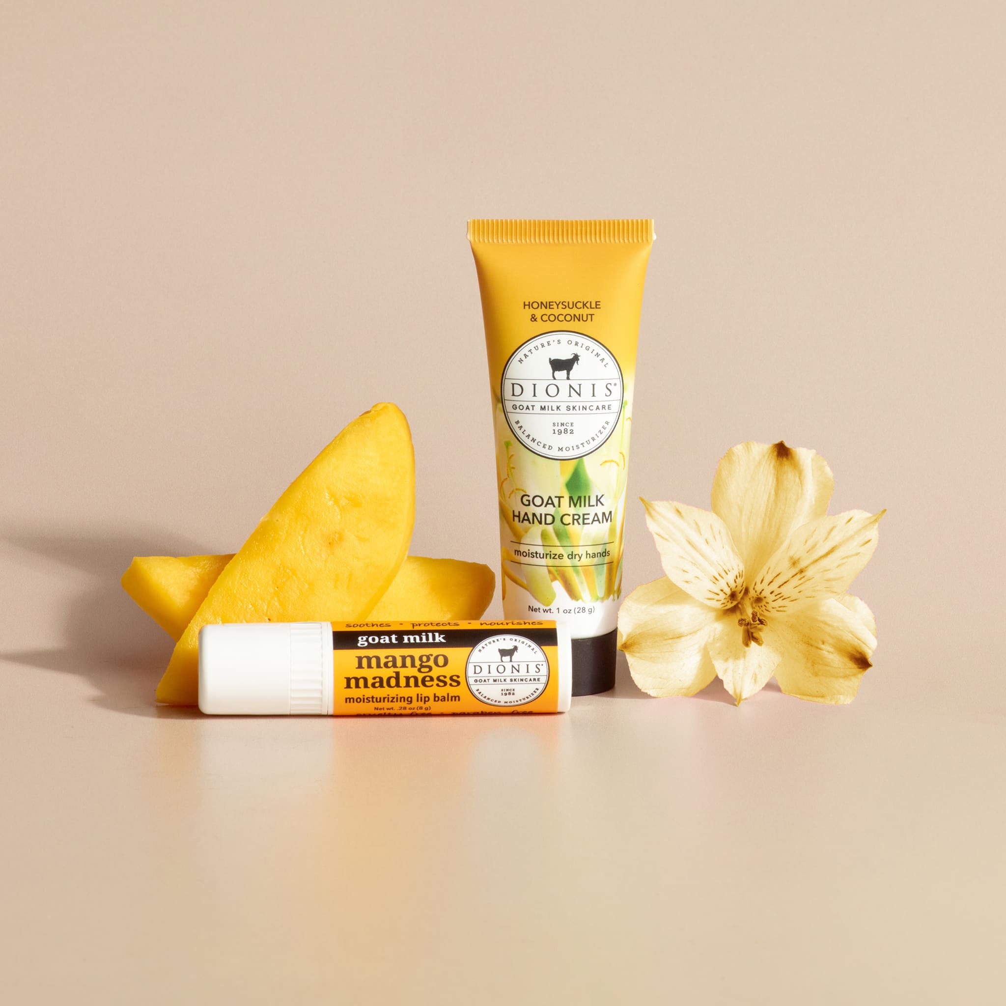 Dionis Goat Milk Skincare Honeysuckle & Coconut hand cream and Mango Lip Balm next to fragrance props of sliced mango and a honeysuckle flower, against a plain beige background