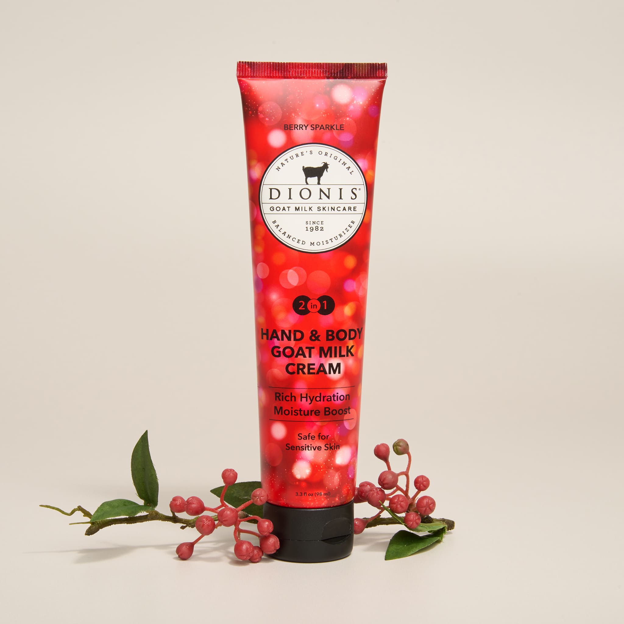 A squeeze tube of Dionis Hand & Body Goat Milk Cream in Berry Sparkle fragrance, stands upright on a light beige background, styled with small red berries and green leaves at the base. The tube is red with light spots to show the sparkle, and a black cap.