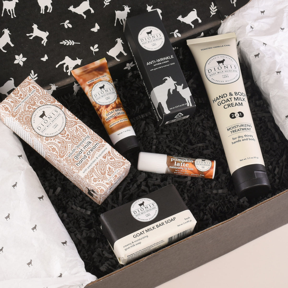 A curated Dioni's Gift Box set containing multiple skincare products including Goat Milk Bar Soap, Hand & Body Goat Milk Cream, Anti-Wrinkle Cream, and Pumpkin Latte lip balm, arranged in black crinkle paper inside a cardboard box with branded tissue paper.