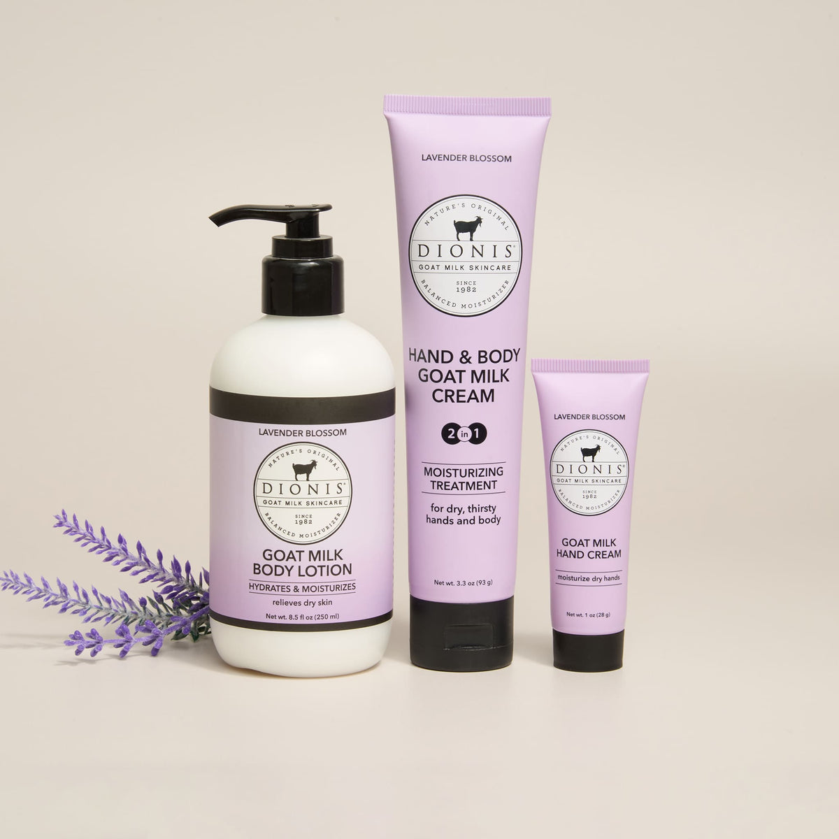 Three Dionis goat milk body care products in Lavender Blossom fragrance arranged on a light beige background: a white pump bottle of body lotion, a lavender tube of hand and body goat milk cream, and a smaller lavender squeeze tube of hand cream, with lavender sprigs at the left.
