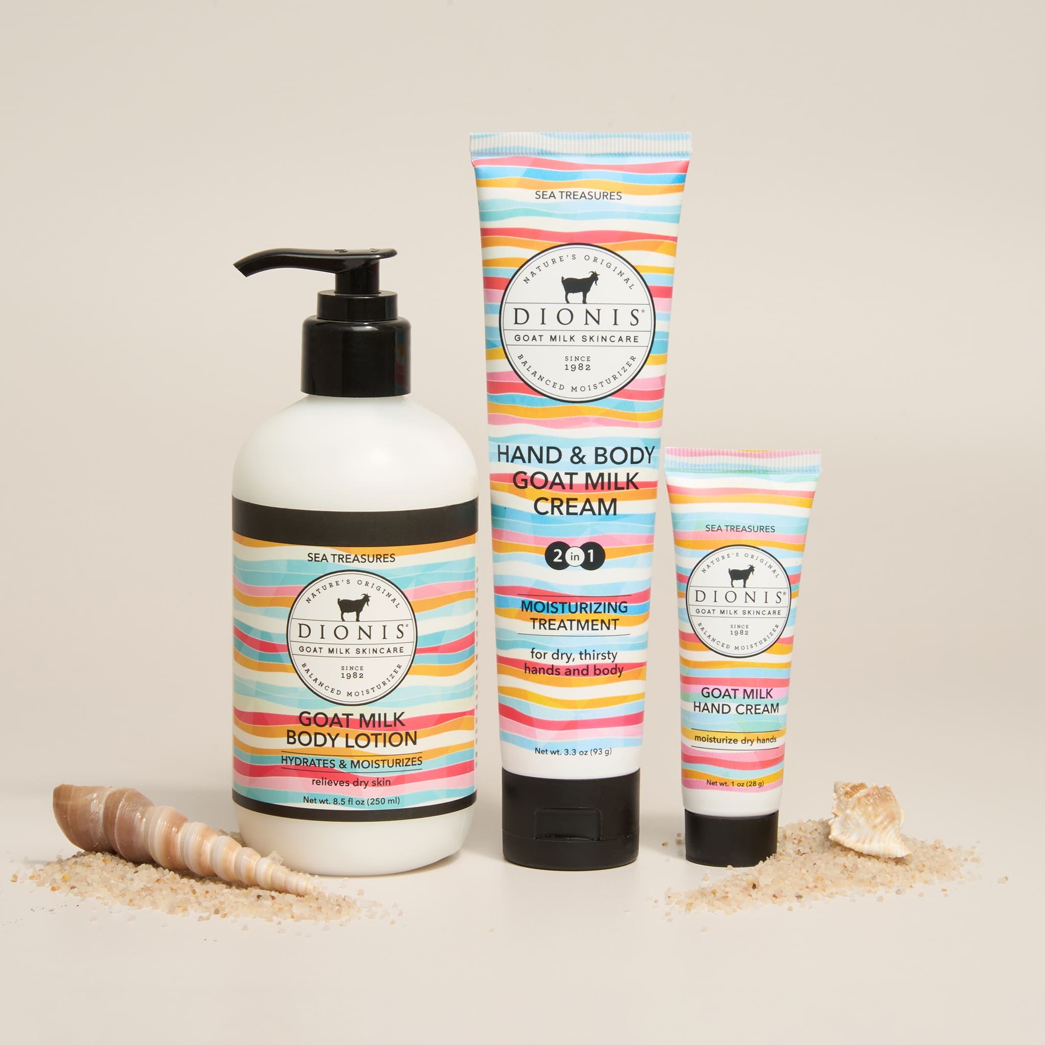 Three Dionis Sea Treasures goat milk skincare products in Sea Treasures Fragrance arranged on a light beige background with scattered seashells: a large pump bottle of goat milk body lotion, a tall tube of hand & body cream, and a smaller tube of hand cream. The labels have colorful horizontal stripes in pink, blue, yellow, and white.