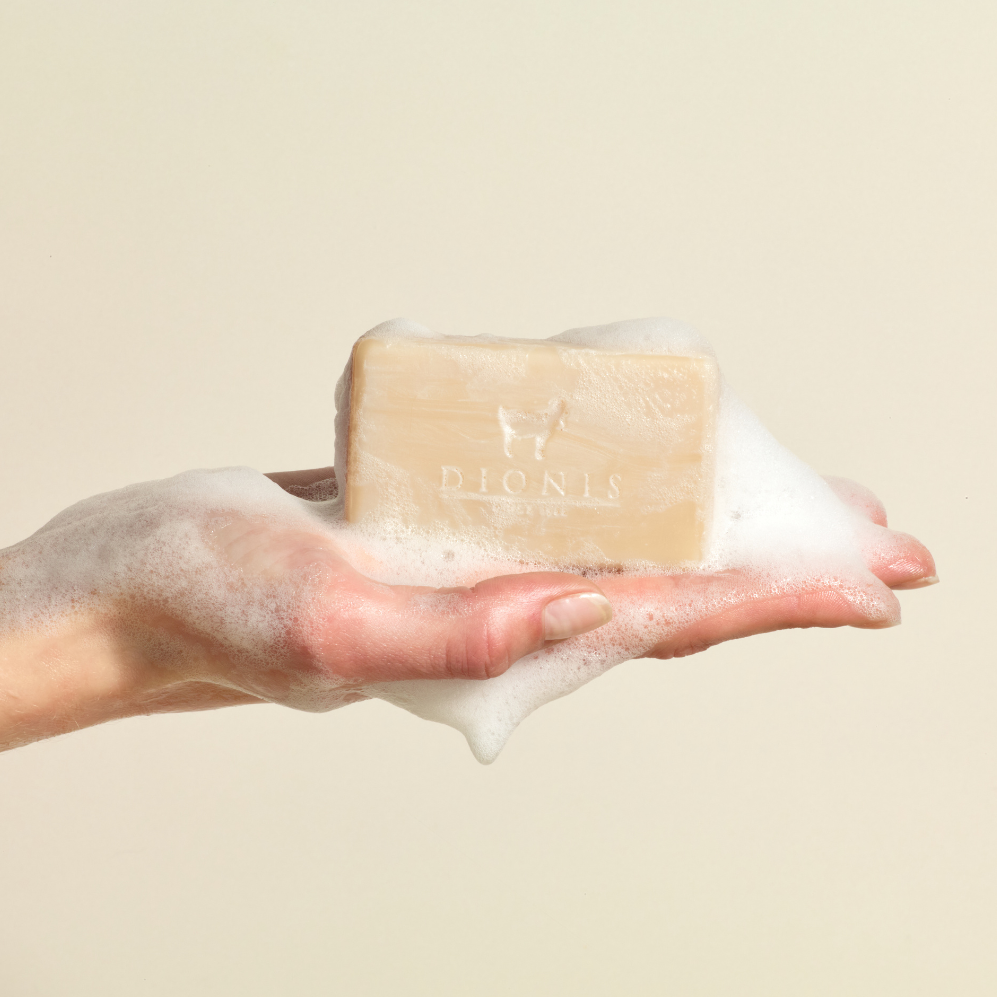 A lathered hand holding a soapy Dionis Bar Soap against a beige background