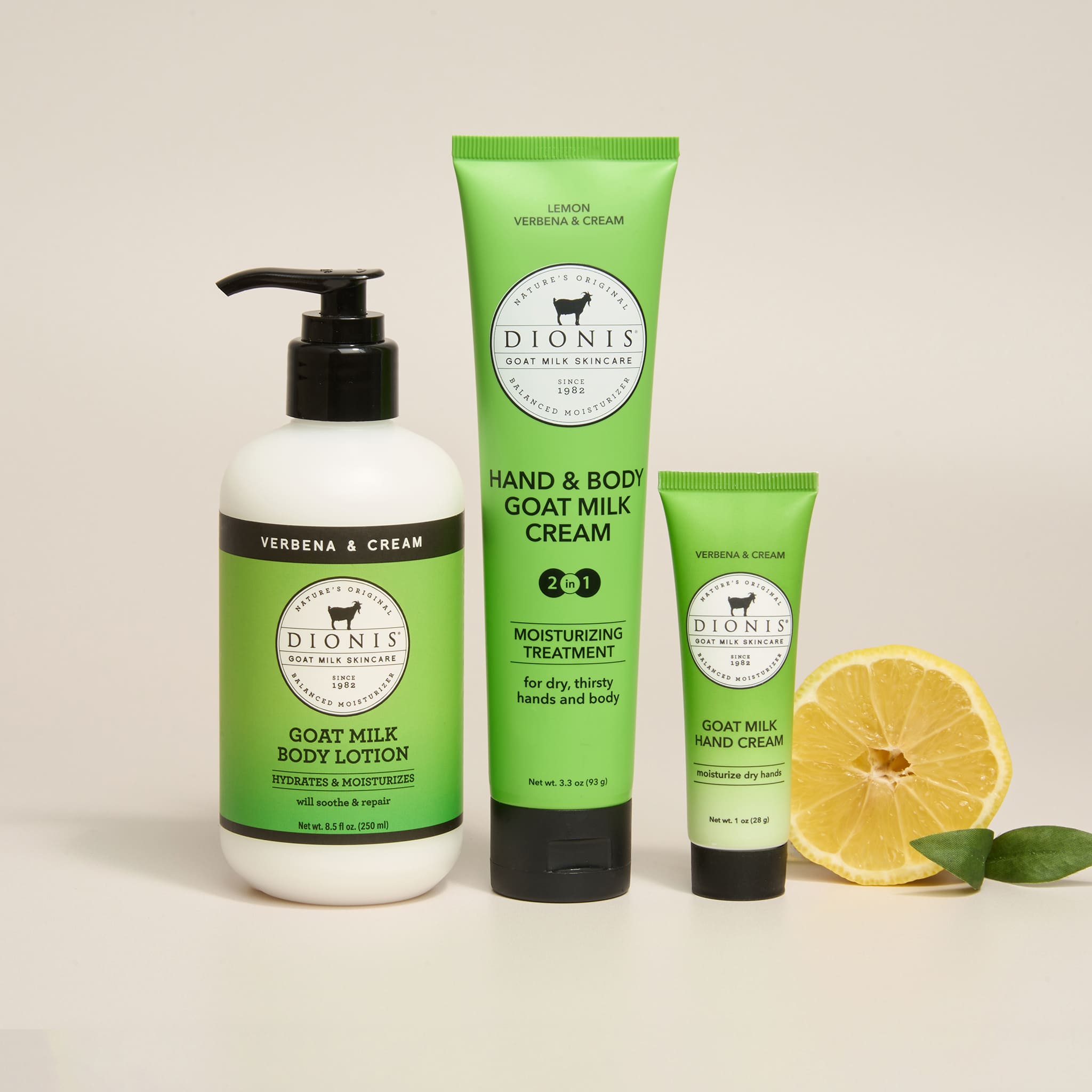 Three Dionis Goat Milk Body Care products in Lemon Verbena fragrance arranged on a light beige background: a white pump bottle of Verbena & Cream body lotion, a green tube of Hand & Body Goat Milk Cream, and a smaller green tube of Goat Milk Hand Cream, with a sliced lemon and green leaves beside them.
