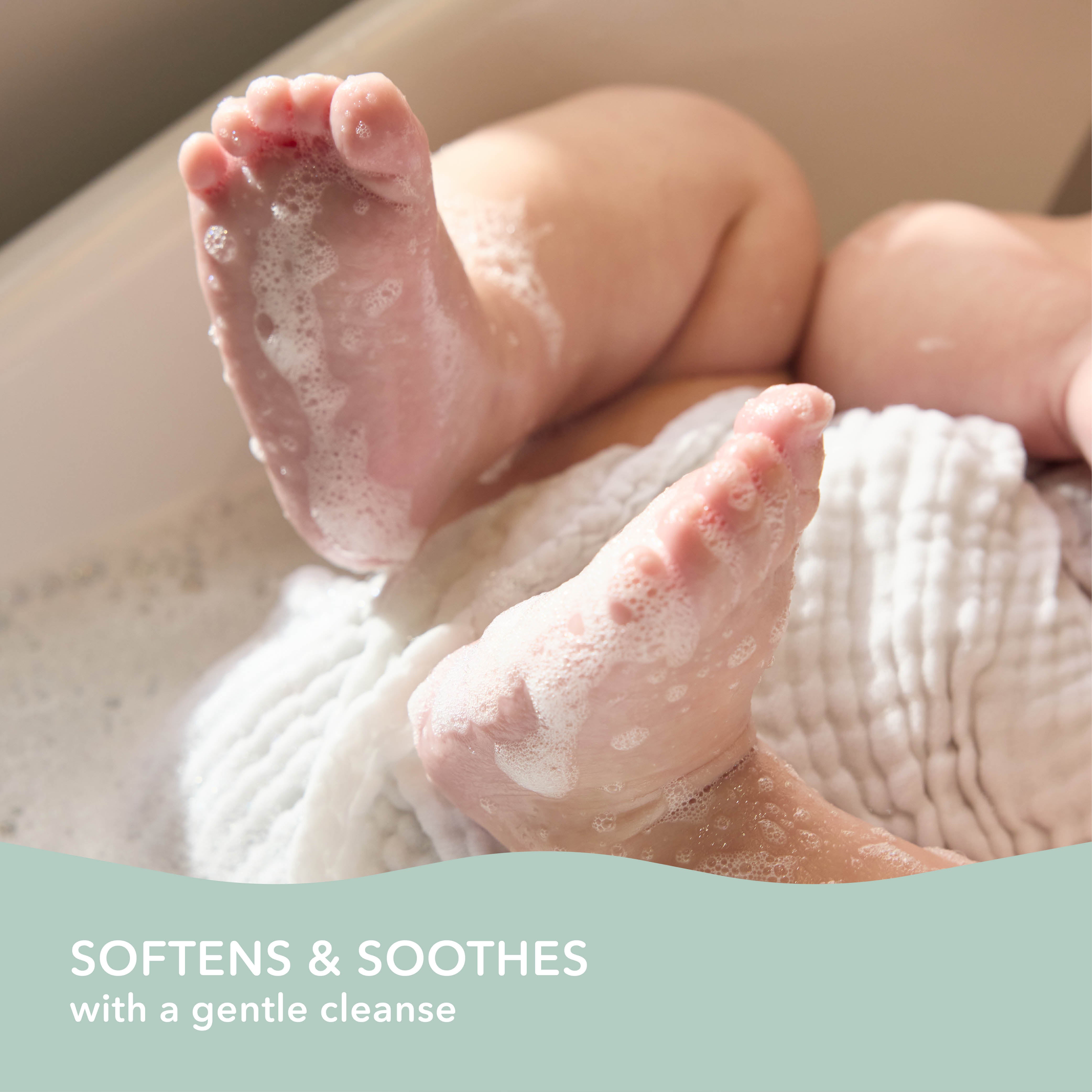 Close-up of a baby's feet and legs wrapped in a soft white towel, with soap lather on the skin in a bath setting. A pale teal banner at the bottom reads 'SOFTENS & SOOTHES with a gentle cleanse'.