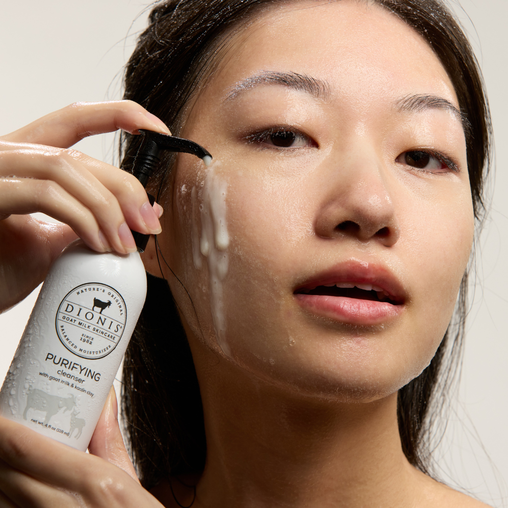 Asian woman holding a bottle of Dionis Goat Milk Skincare Purifying Cleanser in her hands and pumping the cleanser onto her face