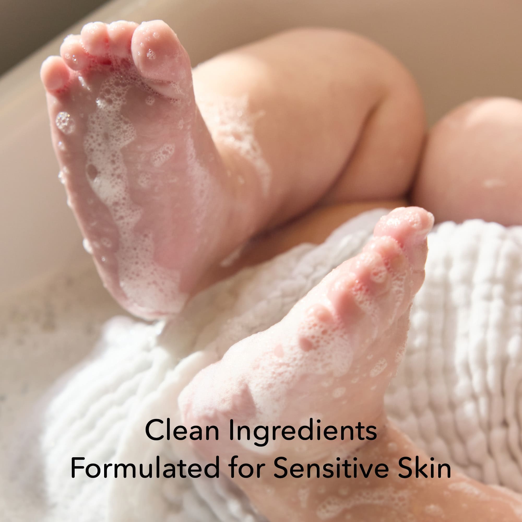 Close-up of a baby's bare feet covered in white suds, resting on a white textured wash cloth. Text at the bottom reads 'Clean Ingredients Formulated for Sensitive Skin'.