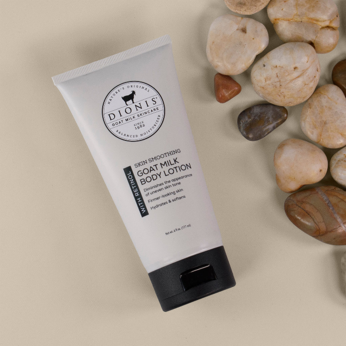 A tube of Dionis Goat Milk Skincare goat milk body lotion with retinol lying on a beige surface iwth rocks and stones as props.