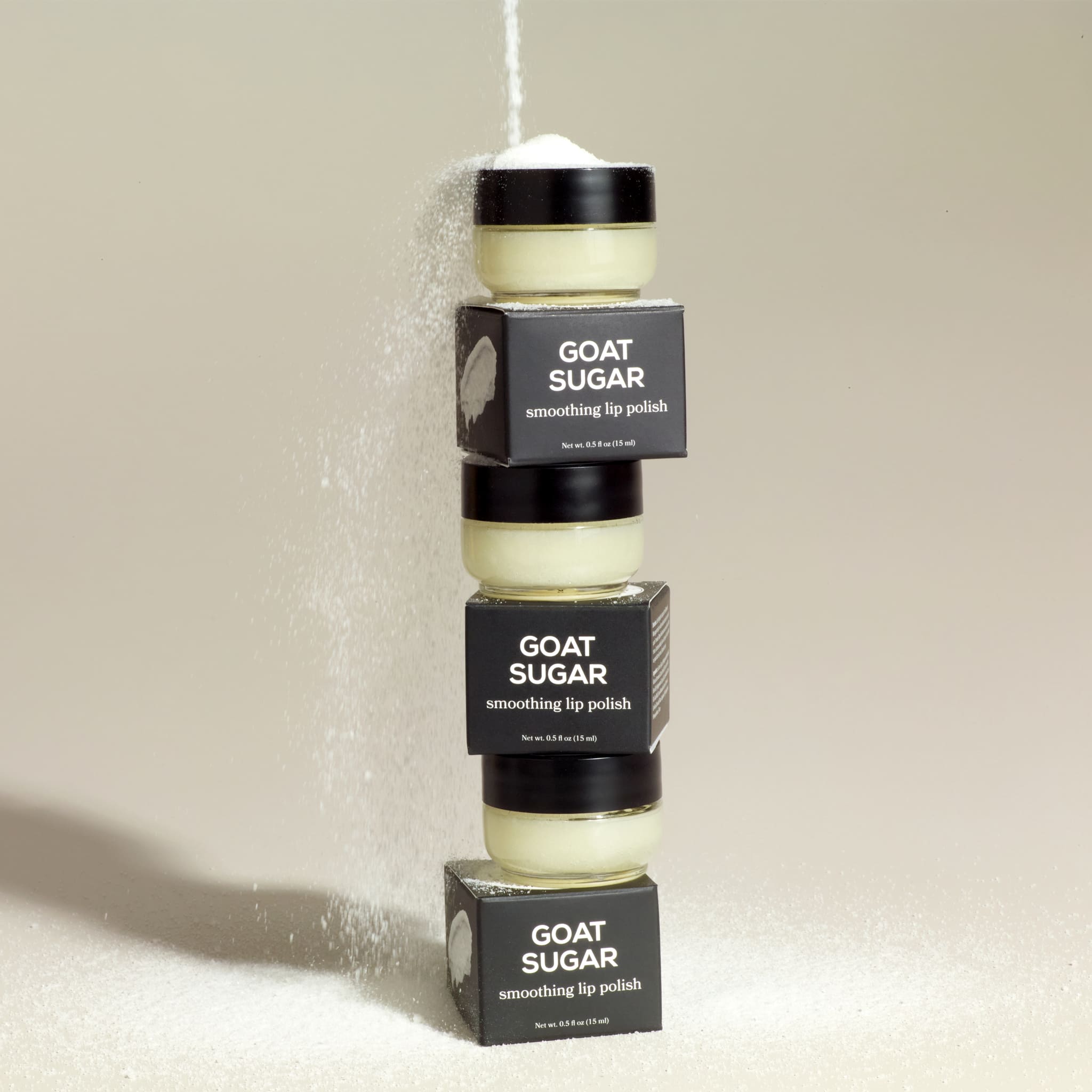 A stack of 3 Dionis Goat sugar lip polishes with their boxes under a stream of sugar poured from the top