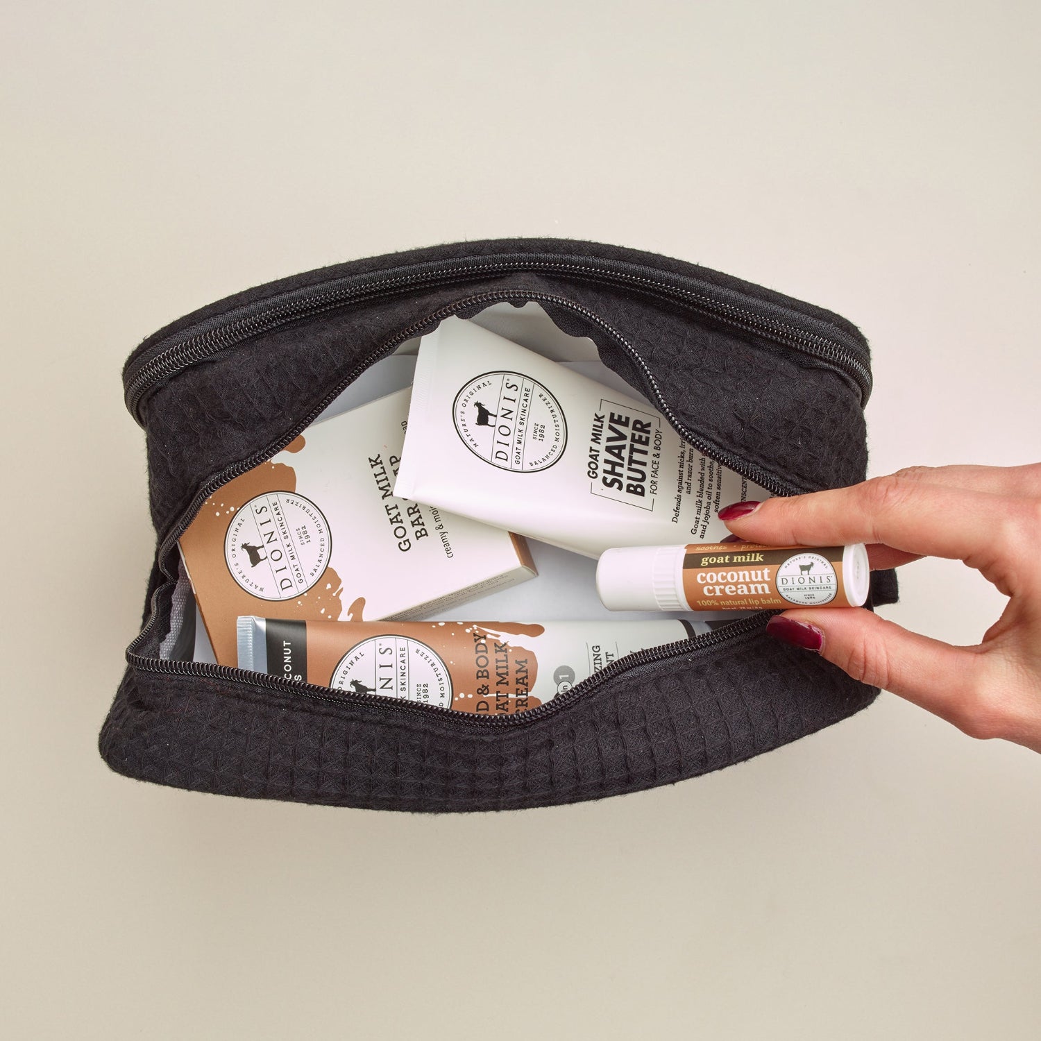 Dionis large vanity bag with Dionis Goat Milk Skincare products inside and a hand pulling an item out, all against a beige background