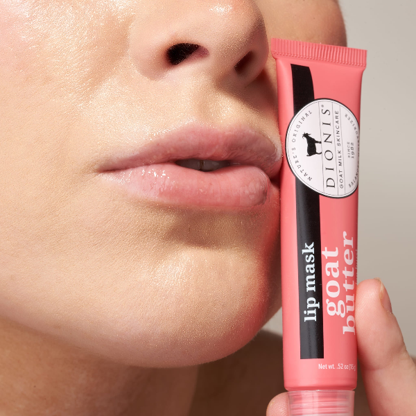 Woman holding a tube of Dionis Goat Butter Lip Mask in Watermelon Burst flavor against her face directly next to her lips which are pouted.