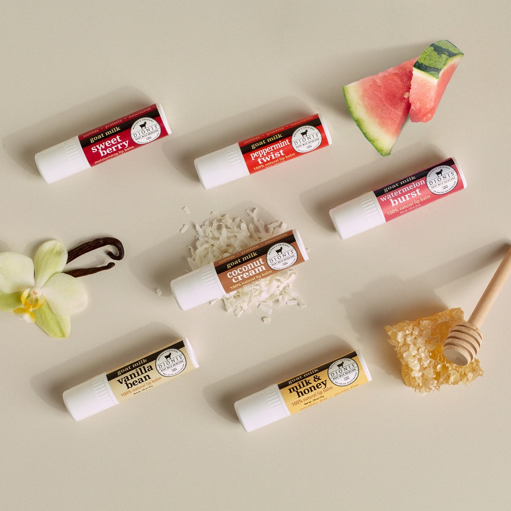 Dionis Goat Milk Lip Balms spread out against a beige background with accompanying props to represent fragrances like coconut, watermelon and vanilla