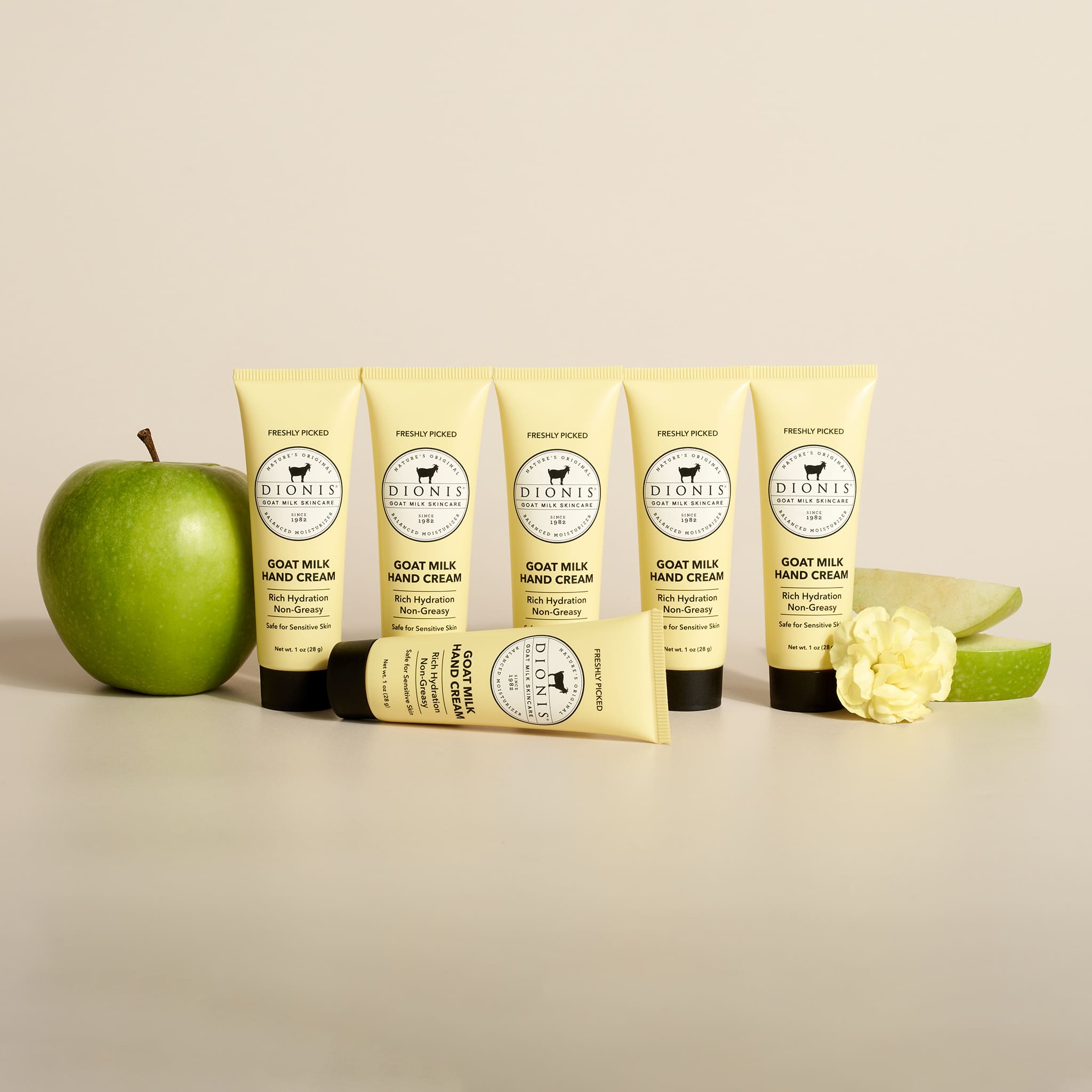 6 tubes of Dionis Goat Milk Skincare 1 oz Freshly Picked Goat Milk Hand Cream with apple and flower props against a plain beige background.
