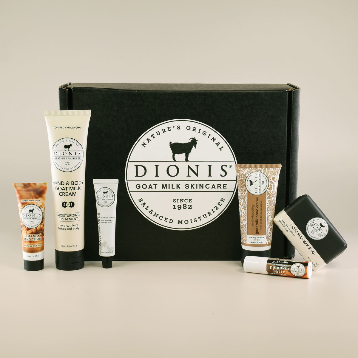Dionis Goat Milk Skincare gift set featuring a black cardboard box with the brand logo, surrounded by various skincare products including hand and body creams, anti-wrinkle treatment, lip balm, and a soap bar, arranged on a light beige background.