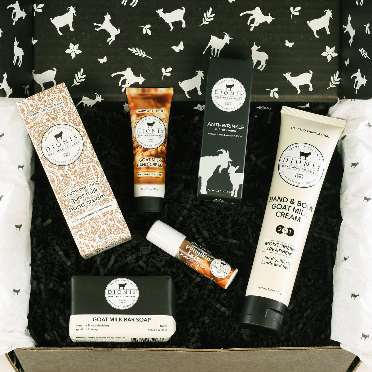 A curated goat milk skincare gift box from Dionis, featuring a hand cream, hand & body lotion, anti-wrinkle cream, lip balm, and goat milk bar soap arranged in black shredded paper inside a brown cardboard box with white patterned tissue paper.