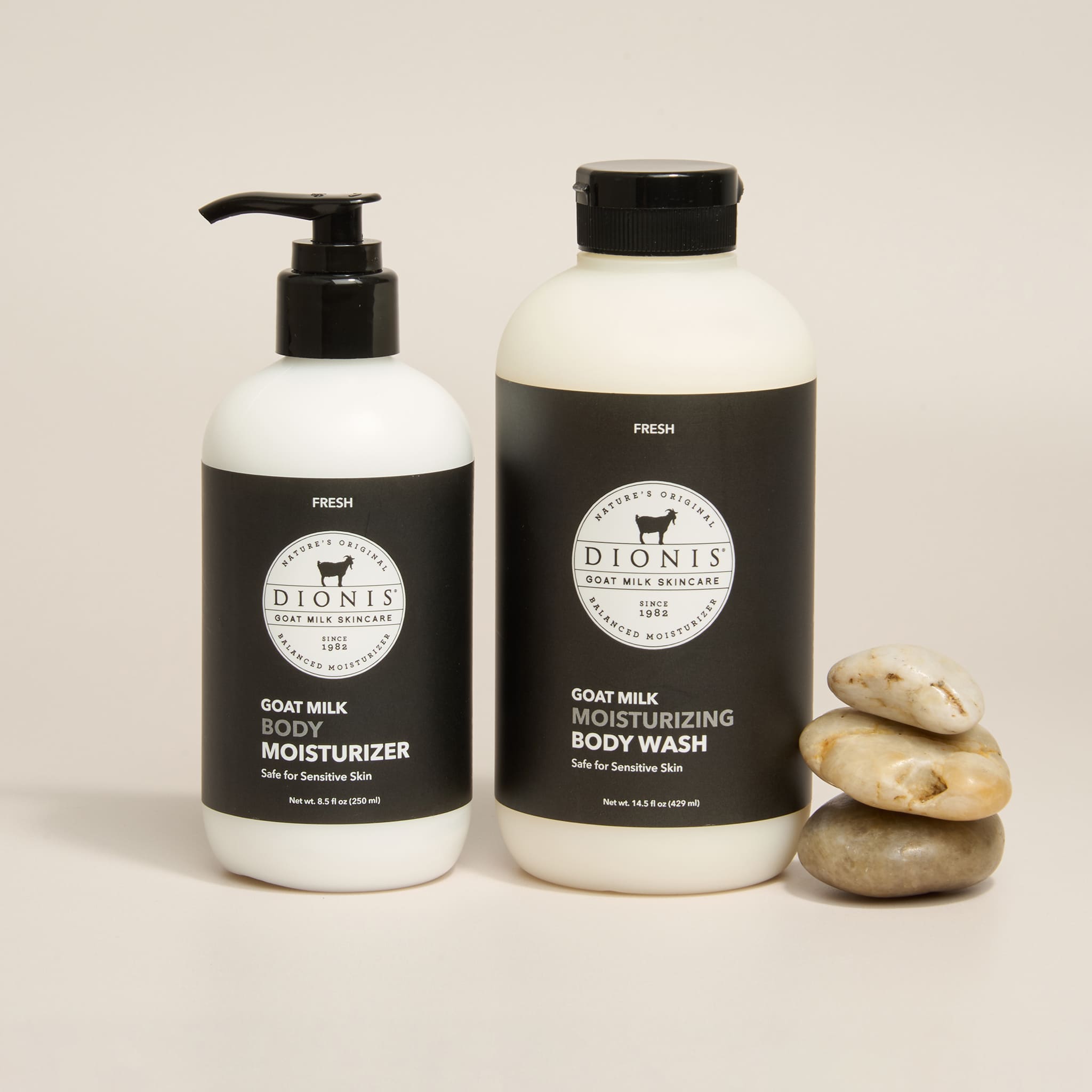 Two bottles of Dionis Goat Milk Body Care products on a light beige background: a pump bottle of Goat Milk Body Moisturizer and a cap-screw bottle of Goat Milk Moisturizing Body Wash, with a small stack of smooth stones beside them.