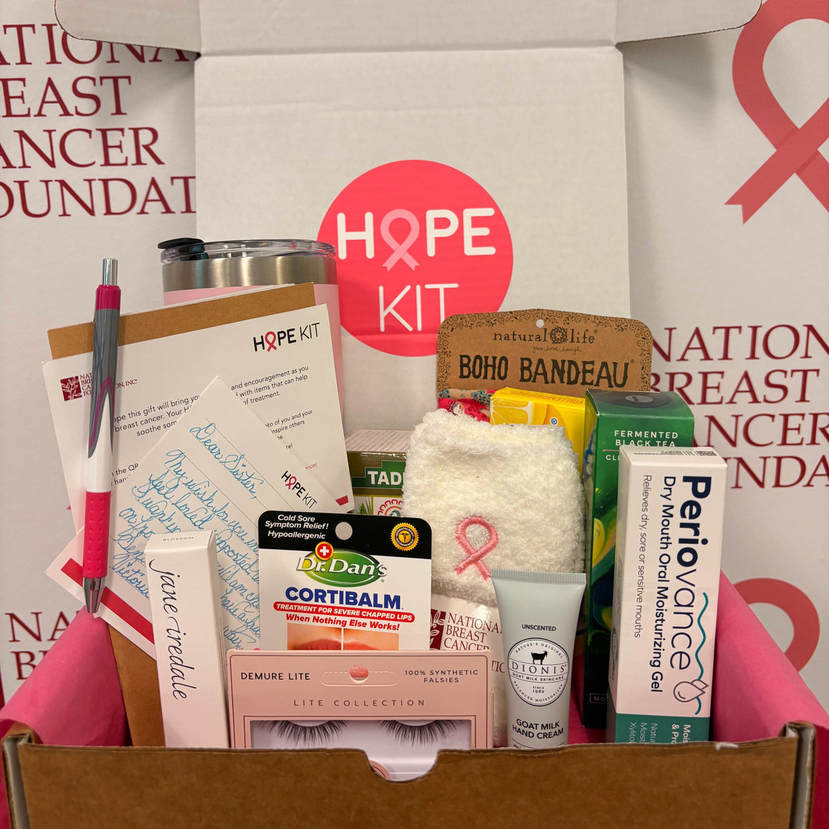 An open cardboard subscription box labeled 'HOPE KIT' with breast cancer awareness ribbon graphics, filled with various wellness and beauty products including a notebook, lip balm, false lashes, and a tube of skin care gel.