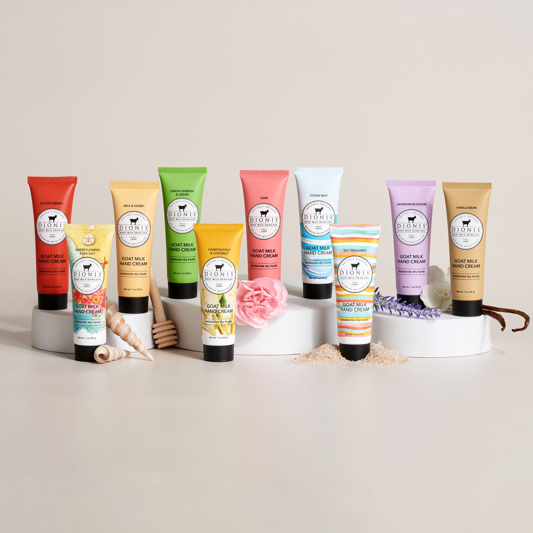 Dionis Goat Milk Hand Creams in a row with various props to indicate the fragrances shown. From left to right, Blood Orange, Water Flowers & Sea Salt, Milk & Honey, Lemon Verbena & Cream, Honeysuckle & Coconut, Love, Ocean Mist, Sea Treasures, Lavender Blossom, Vanilla Bean