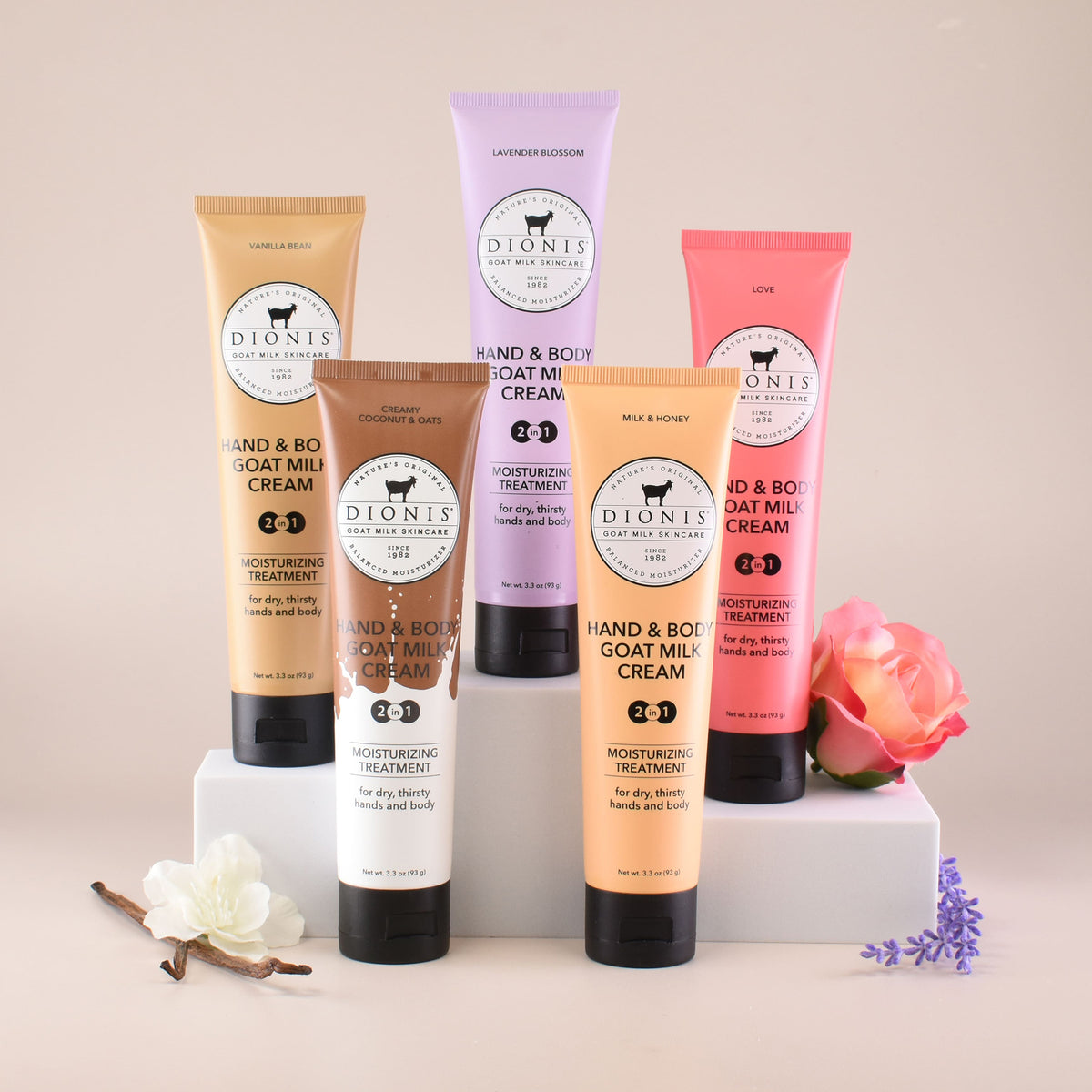 Five tubes of Dionis Hand & Body Goat Milk Cream in assorted fragrances (vanilla bean, creamy coconut & oats, lavender blossom, milk & honey and love) arranged on white pedestals with decorative flowers on a light beige background.