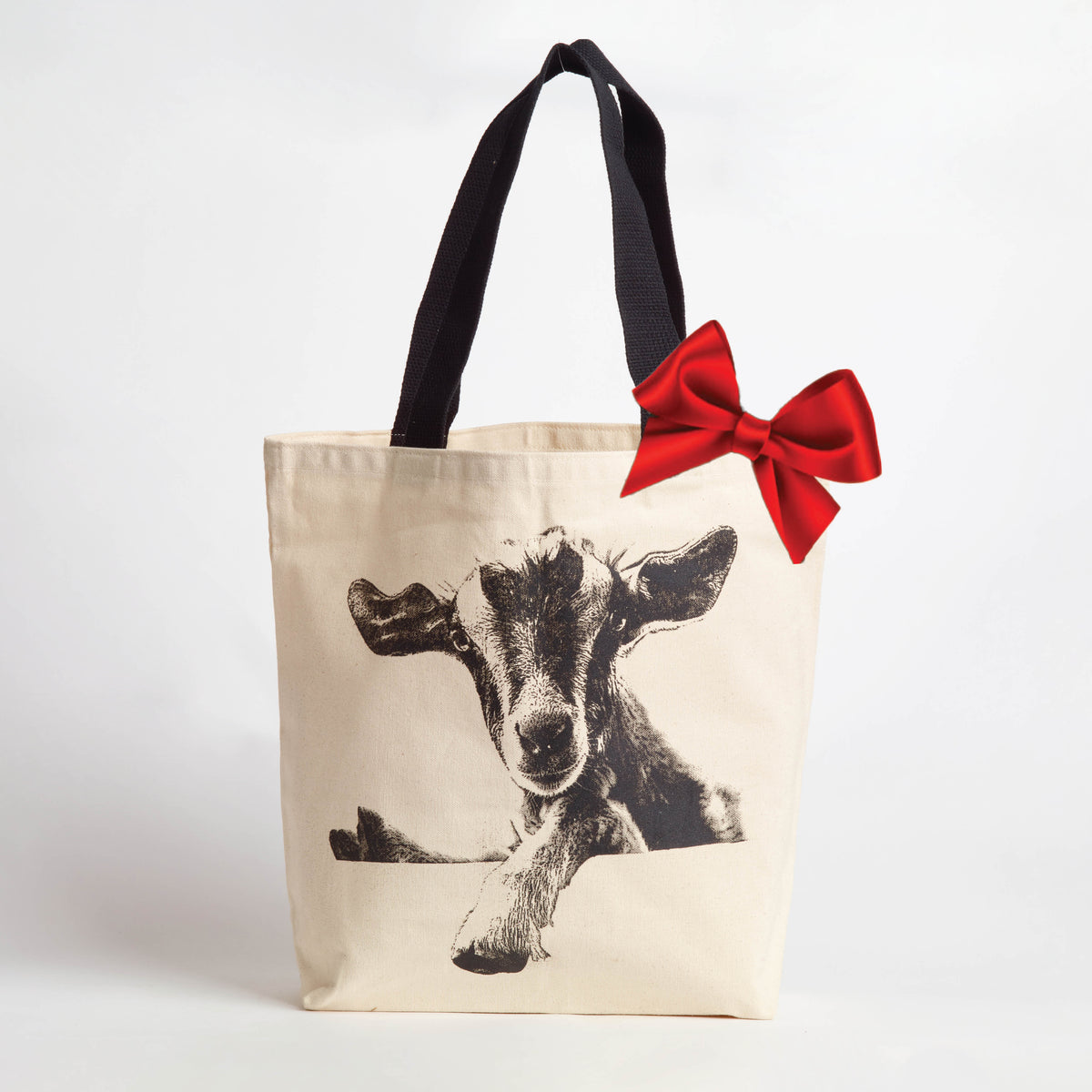 A cream canvas tote bag with black handles, featuring a black graphic print of a goat and a large red satin bow on the side, set against a plain white background.