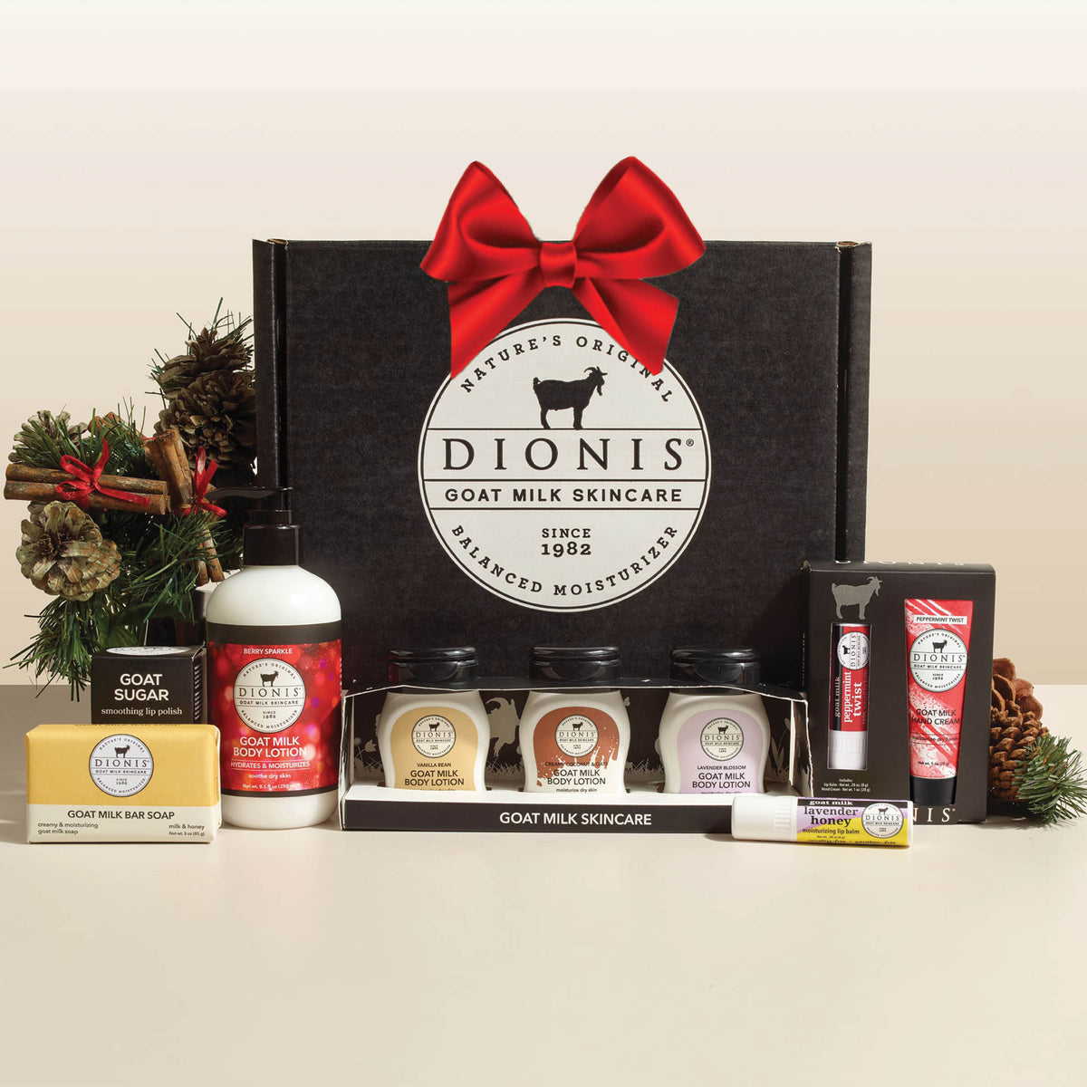 Dionis Goat Milk Skincare gift set in a black box with a red bow, displayed with various skincare products including hand cream, sugar scrub, body lotions, bar soap, and lip balm on a light beige background.