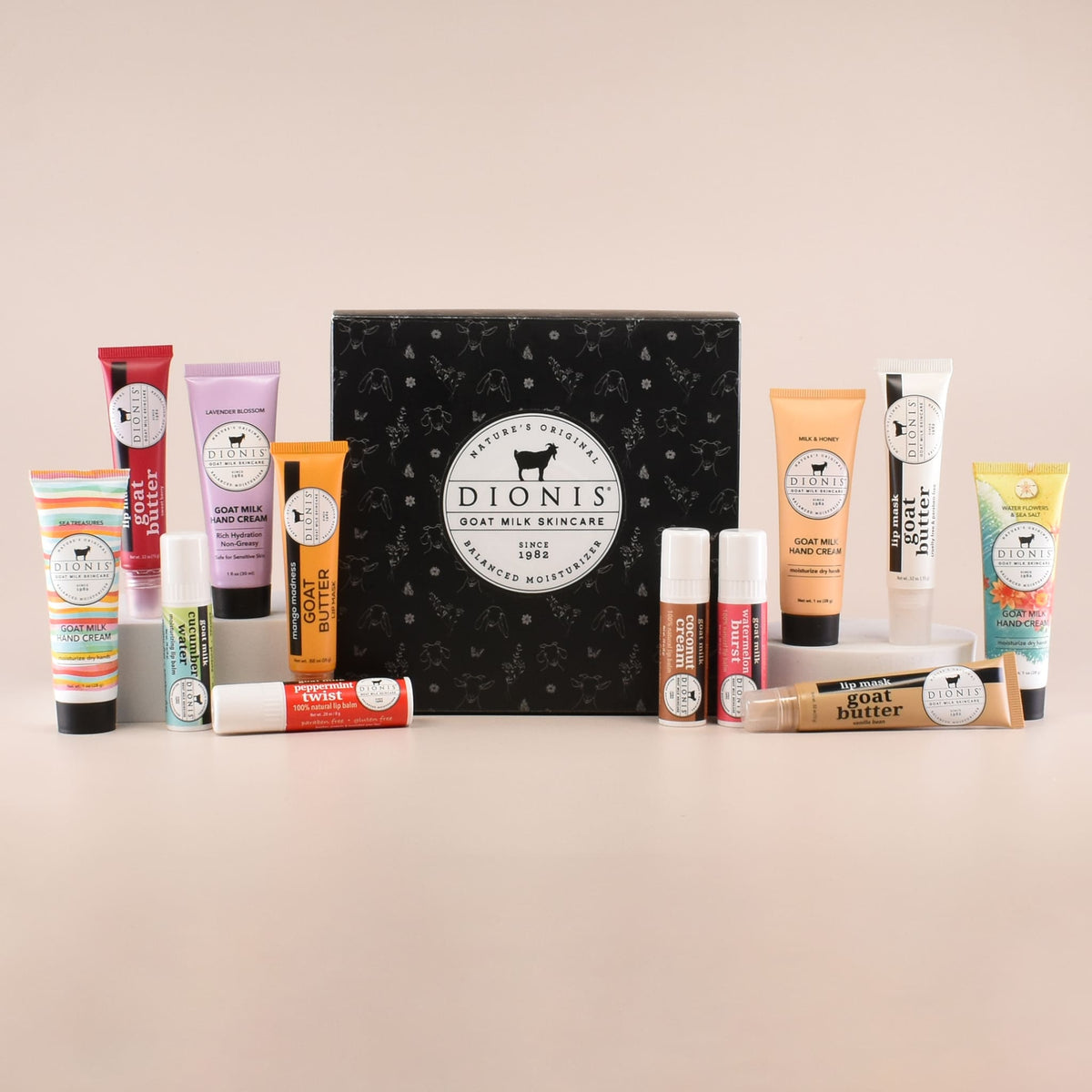 A Dionis Goat Milk Skincare gift set arranged on a light beige background, featuring a black patterned box surrounded by various hand cream tubes, lip balms, and a lip masks.