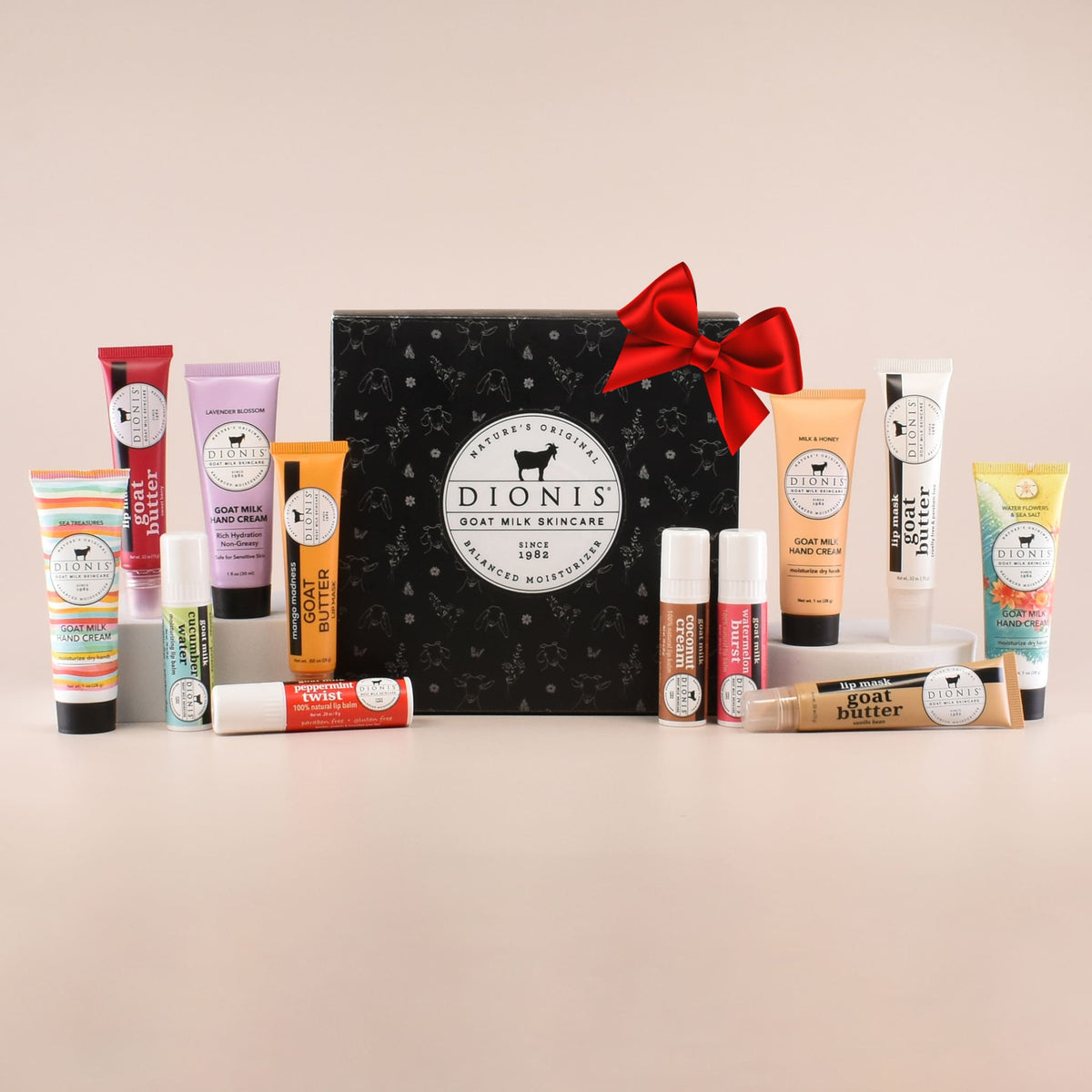 A Dionis Goat Milk Skincare gift set arranged on a light beige background, featuring a black patterned box surrounded by various hand cream tubes, lip balms, and a lip masks. A red bow is on the actual gift box to represent it's a great gift.
