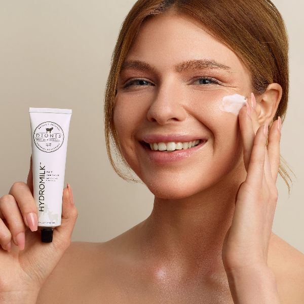 Woman holding a tube of Dionis Goat Milk Skincare Hydromilk 24/7 Moisturizer in one hand and applying the cream to her face with her other hand.