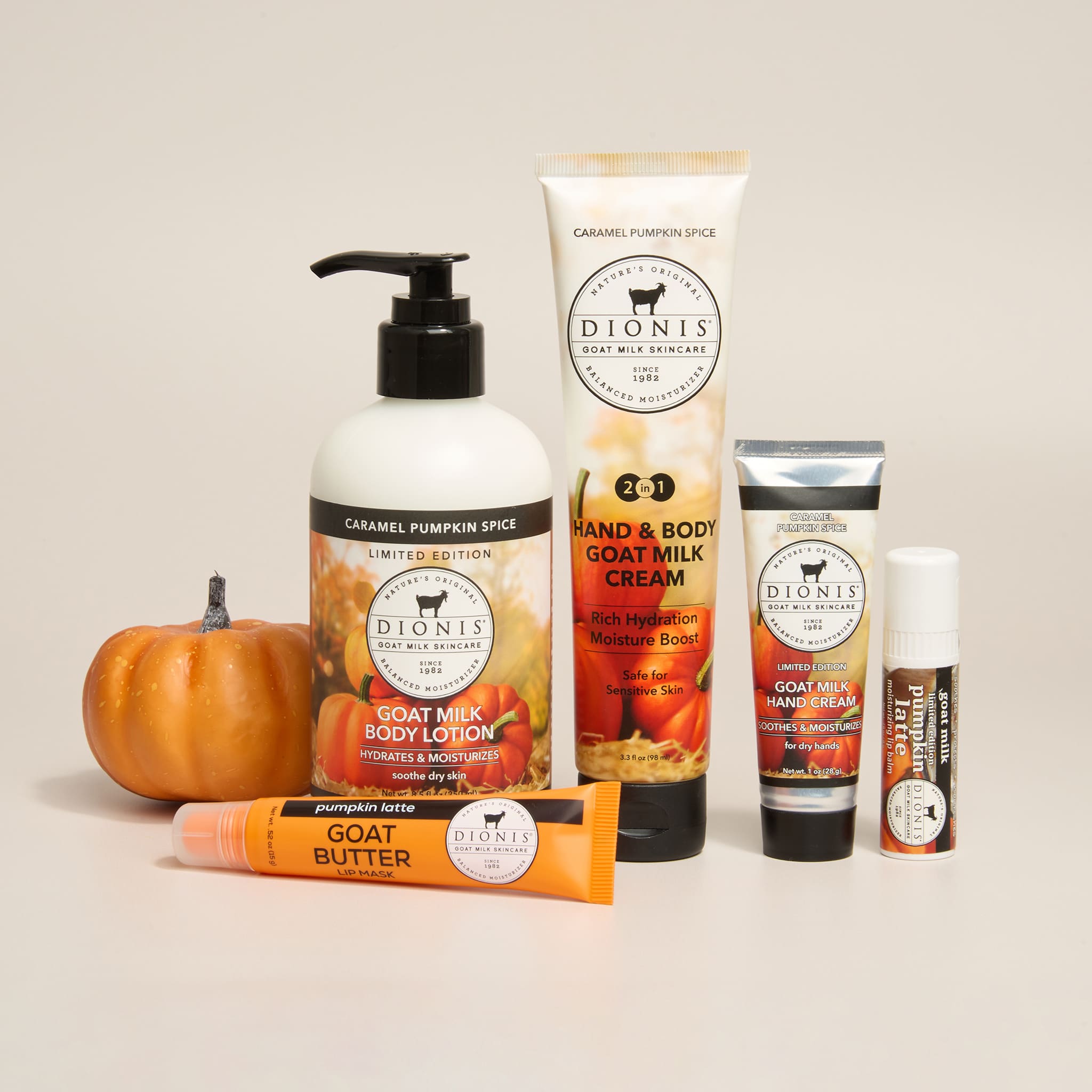 A coordinated set of Pumpkin fragrance Dionis Goat Milk Skincare products arranged on a light warm beige background, including a pump bottle of caramel pumpkin spice body lotion, a hand and body cream tube, a hand and body lotion squeeze tube, a lip balm tube, and a lip mask tube, with a small pumpkin prop at left.