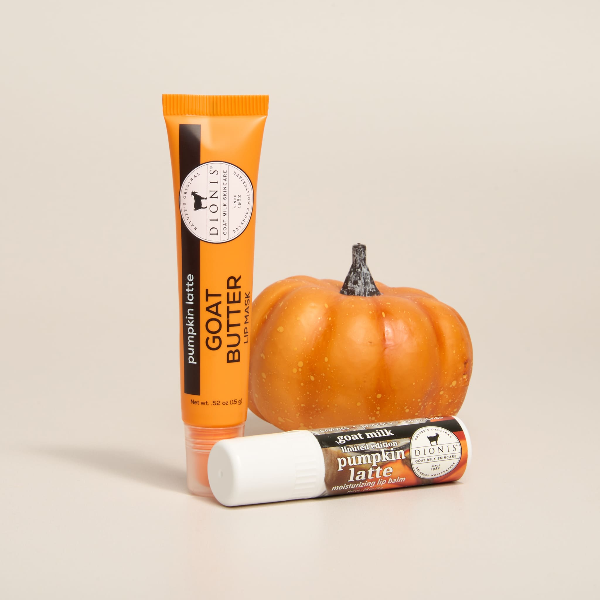 A tube of Dionis Pumpkin Latte Goat Butter Lip Mask and a tube of Pumpkin Latte goat milk lip balm arranged on a light beige background, with a small orange pumpkin placed behind them as a prop.