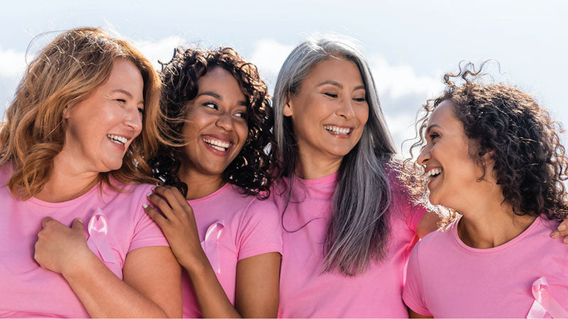 Four women smiling and hugging while wearing matching pink shirts with pink ribbon pins, set against a bright sky background.