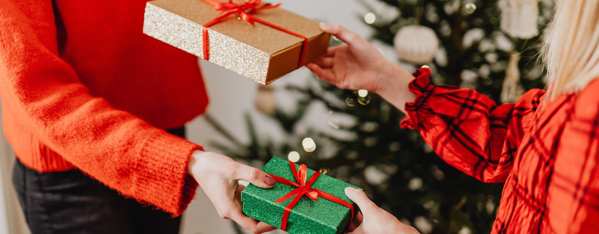 Two people exchanging wrapped holiday gifts in front of a decorated tree. One person holds a gold glitter box with a red ribbon, and the other holds a green box with a red ribbon.