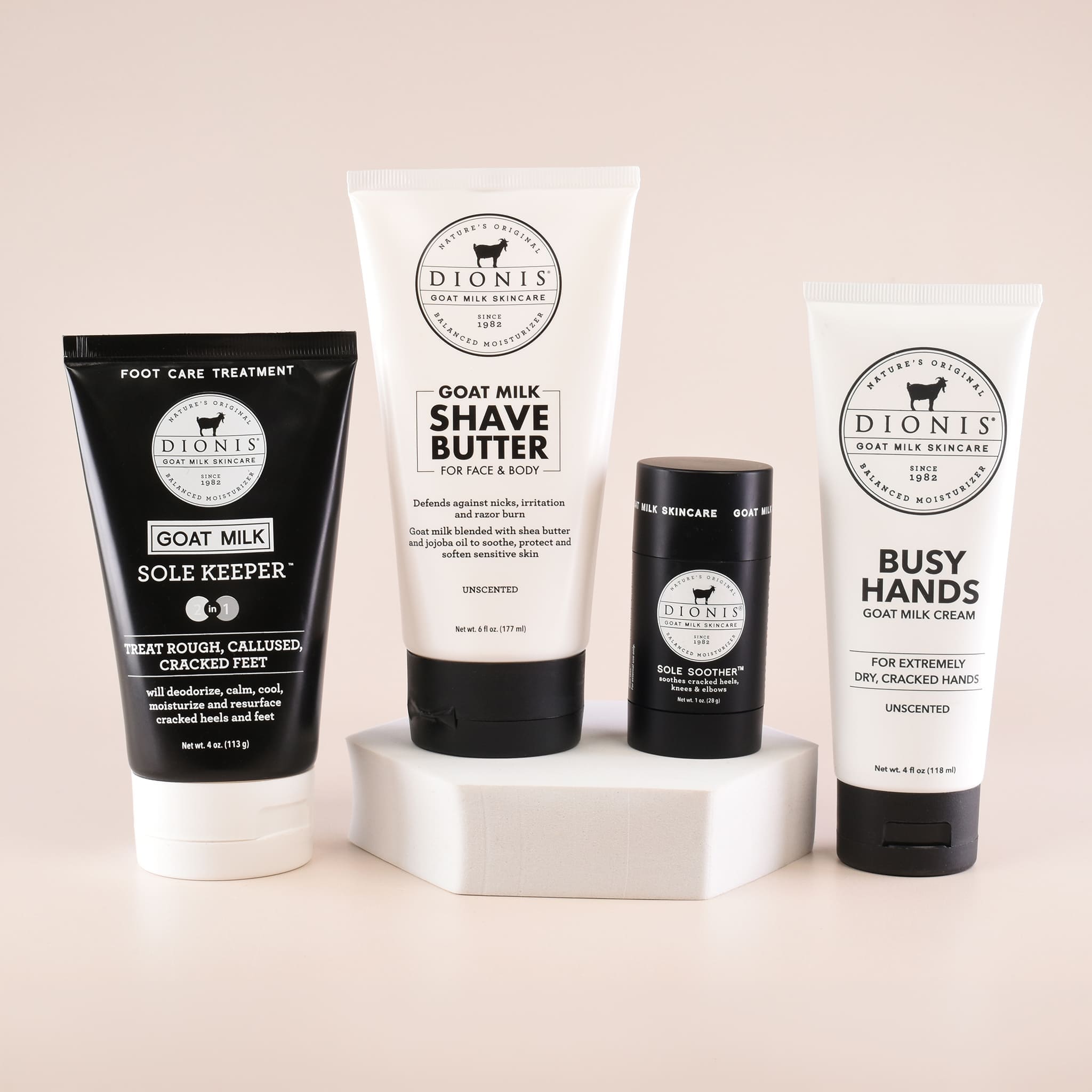 Four Dionis Goat Milk Skincare products arranged on a white pedestal against a warm beige background: a black sole keeper, a white goat milk shave butter tube, a black body balm skincare stick, and a white busy hands cream tube.