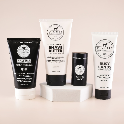 Four Dionis Goat Milk Skincare products arranged on a white pedestal against a warm beige background: a black sole keeper, a white goat milk shave butter tube, a black body balm skincare stick, and a white busy hands cream tube.