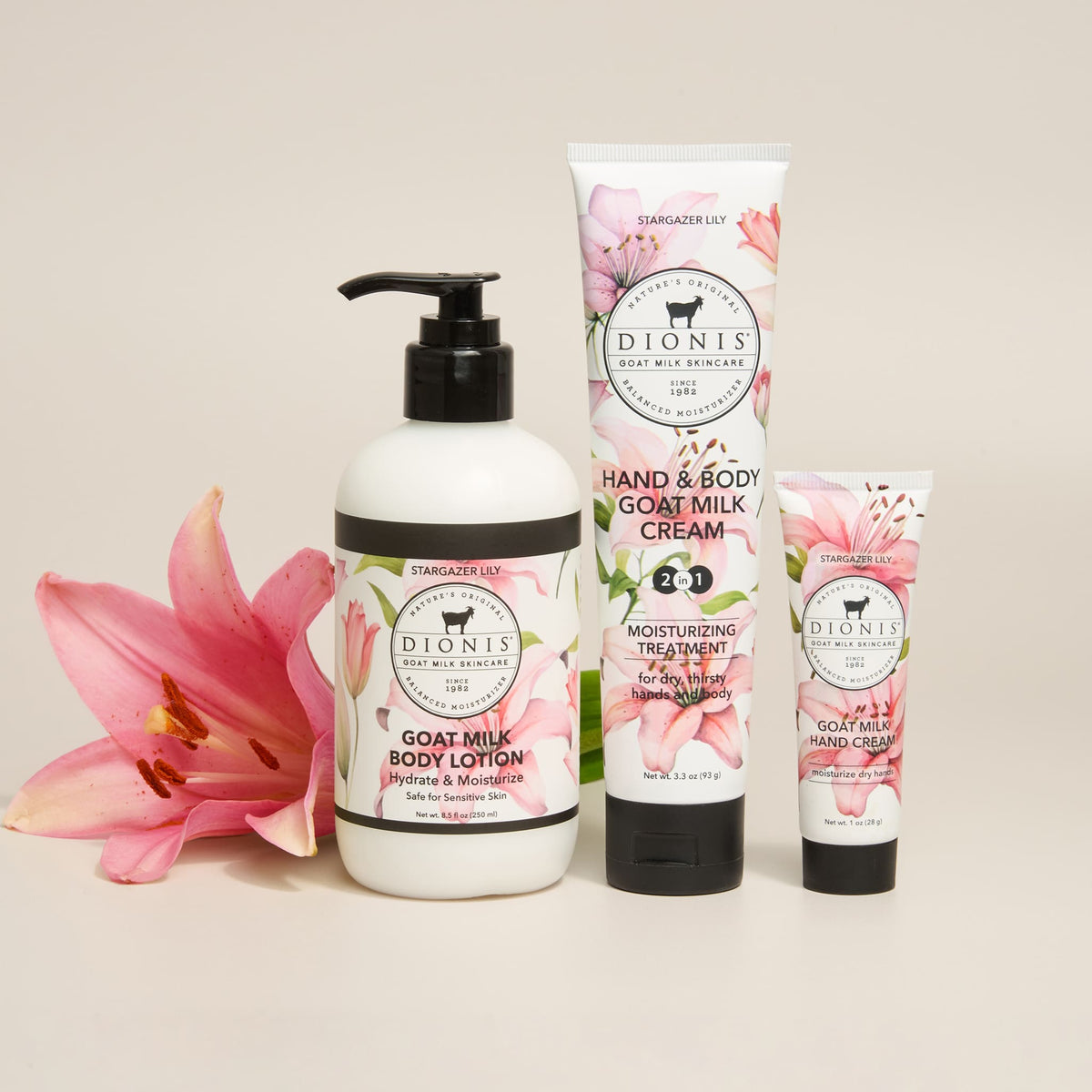 Three Dionis goat milk body care products in Stargazer Lily fragrance arranged on a light beige background with pink lily flowers at the left: a large white pump bottle of body lotion, a tall white tube of hand & body cream, and a smaller white tube of hand cream. The labels are white with pink floral artwork and black text.