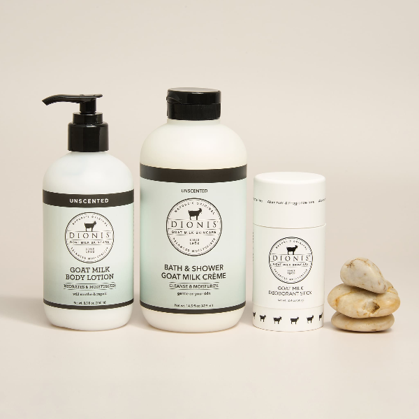 Three white DIONIS goat milk body care products are arranged on a light beige background: a pump bottle of unscented body lotion, a large bottle of unscented bath & shower creme, and a fragrance free deodorant stick, with three natural stones beside them.