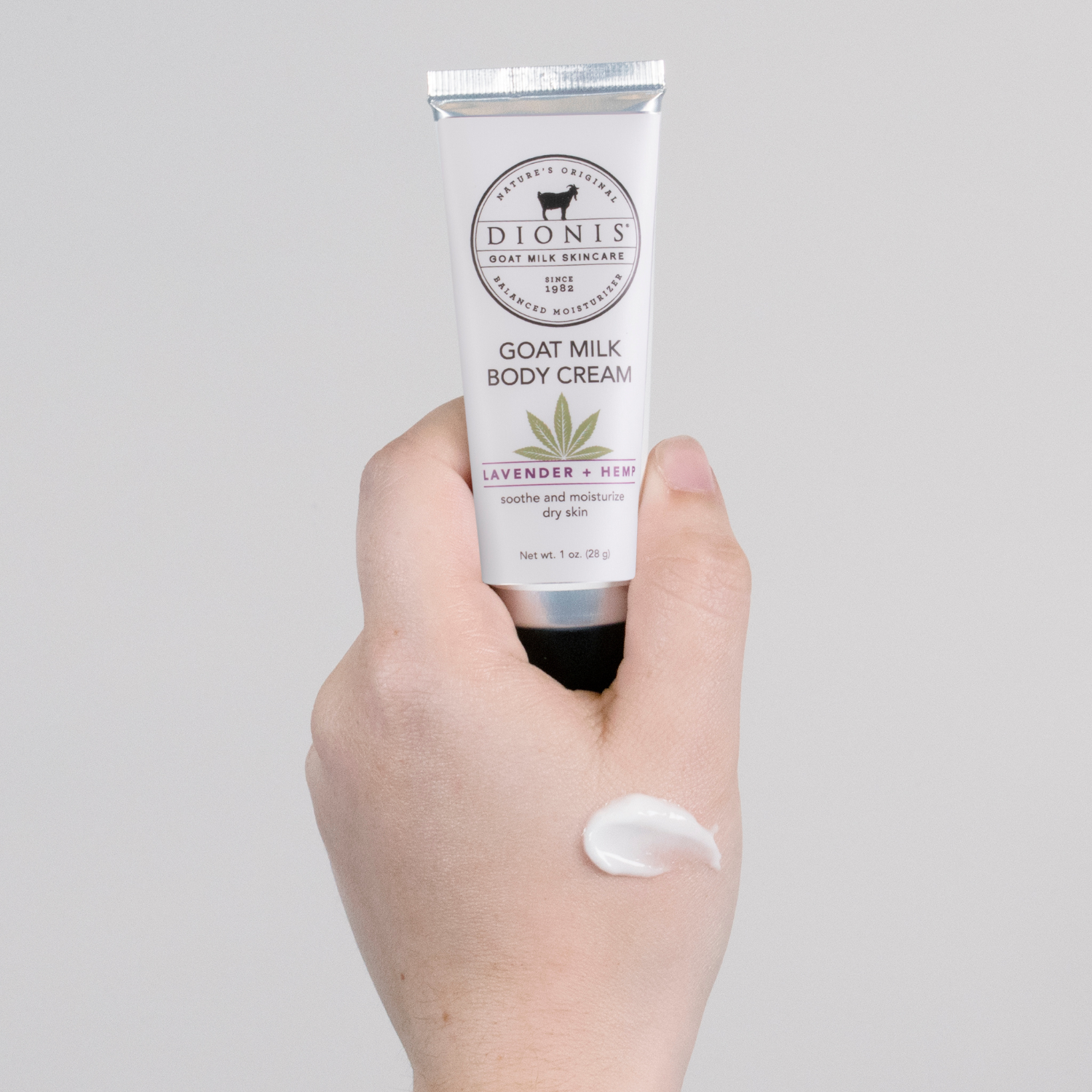 Hand holding a tube of Dionis Goat Milk Lavender + Hemp Body Cream with a visible smear on the hand, against a gray background