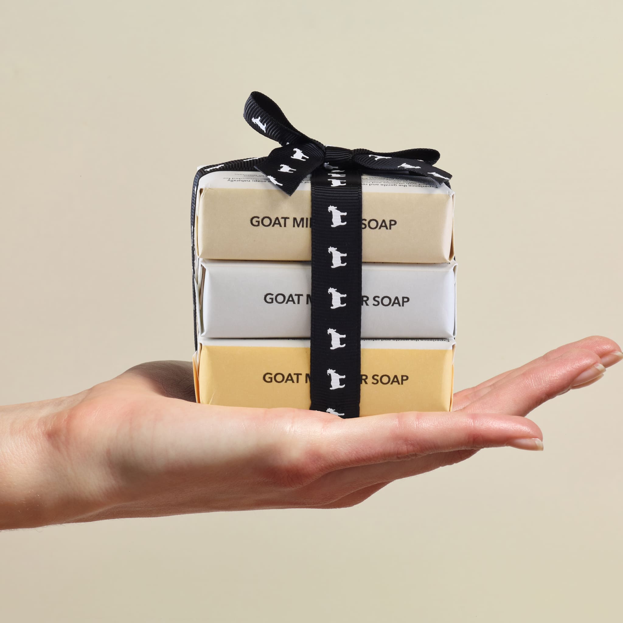 Stack of goat milk soap bars with a black ribbon held in a hand against a beige background