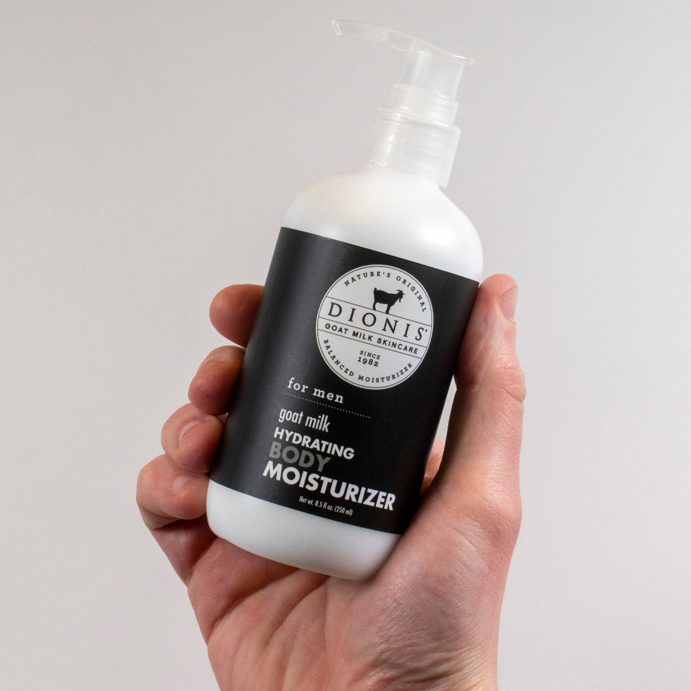 A bottle of Dionis Goat Milk Body Lotion in Men's Fresh scent held by a man's hand on an off-white background.