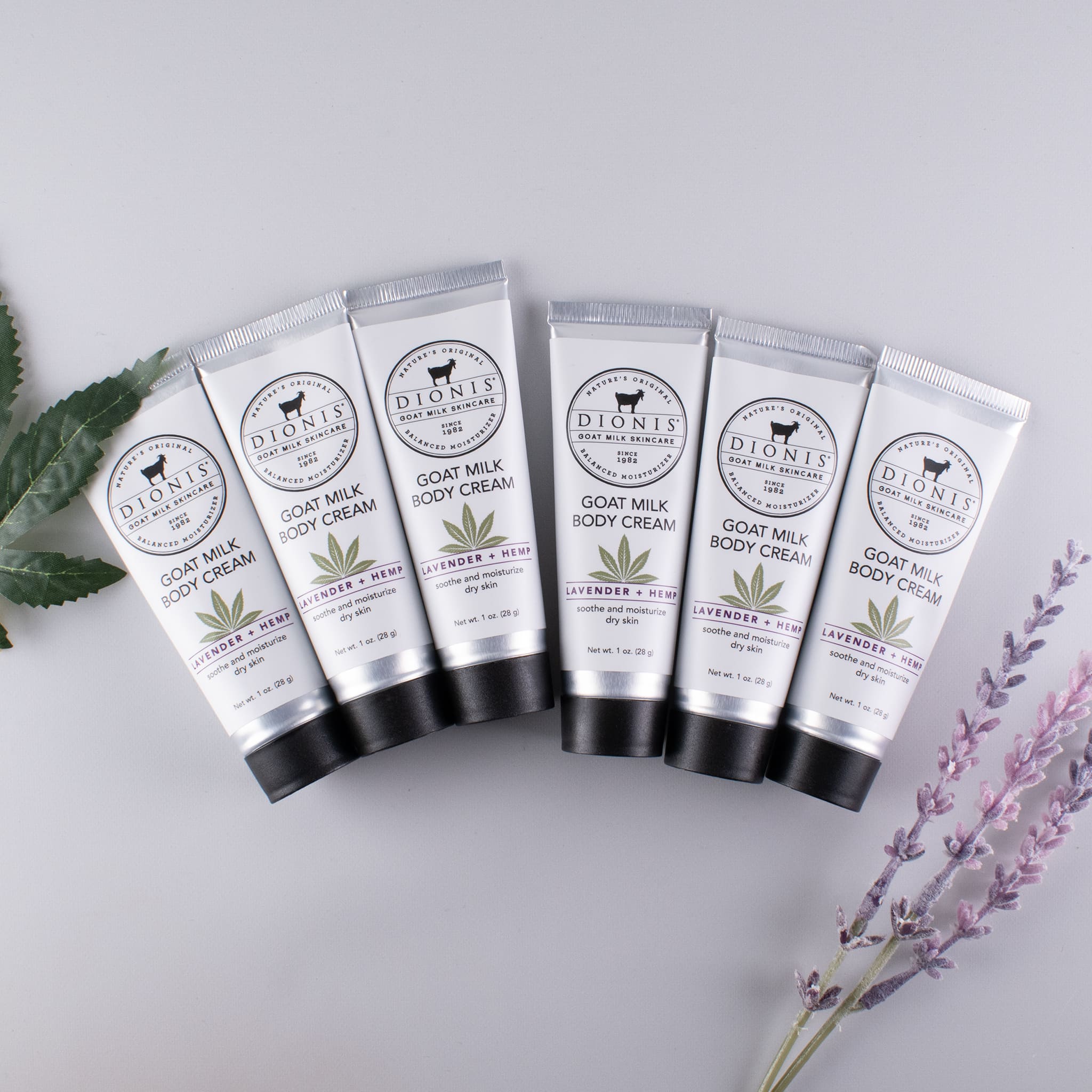 Six tubes of Dionis Lavender + Hemp Goat Milk Hand Cream in a row against a light gray background with a left on the left side and a sprig of lavender on the right side as props representing hemp and lavender.
