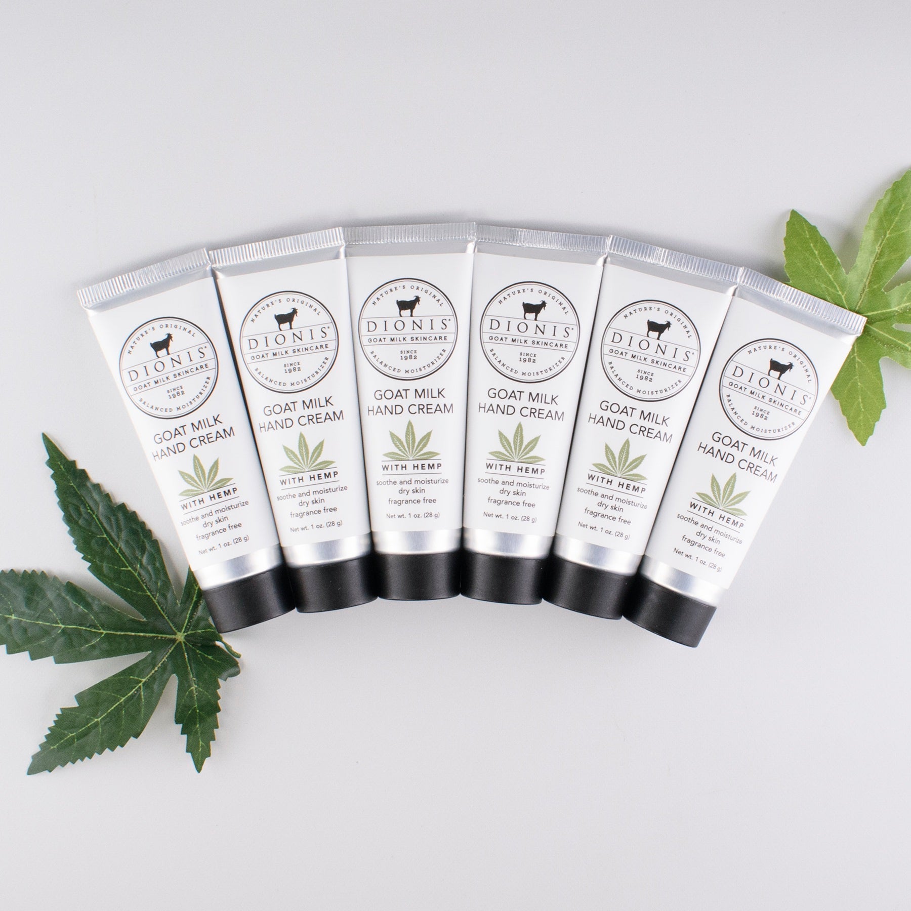 Six Dionis Goat Milk + Hemp 1 oz Hand Cream tubes lined up against a gray background with leaves behind the tubes for props.